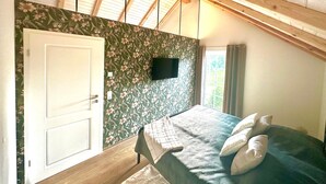Ein Schlafzimmer mit Holzboden, einem Bett mit Kissen, einem an der Wand montierten Fernseher und einem Fenster mit Vorhängen.