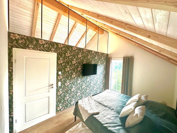 Un dormitorio con techo de madera, una cama con almohadas, un televisor montado en la pared y una ventana con cortinas.
