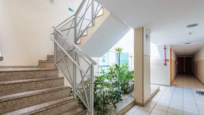 Una escalera con barandillas metálicas que conduce a un piso superior, un pasillo con baldosas y una planta, y una señal de salida de emergencia visible.