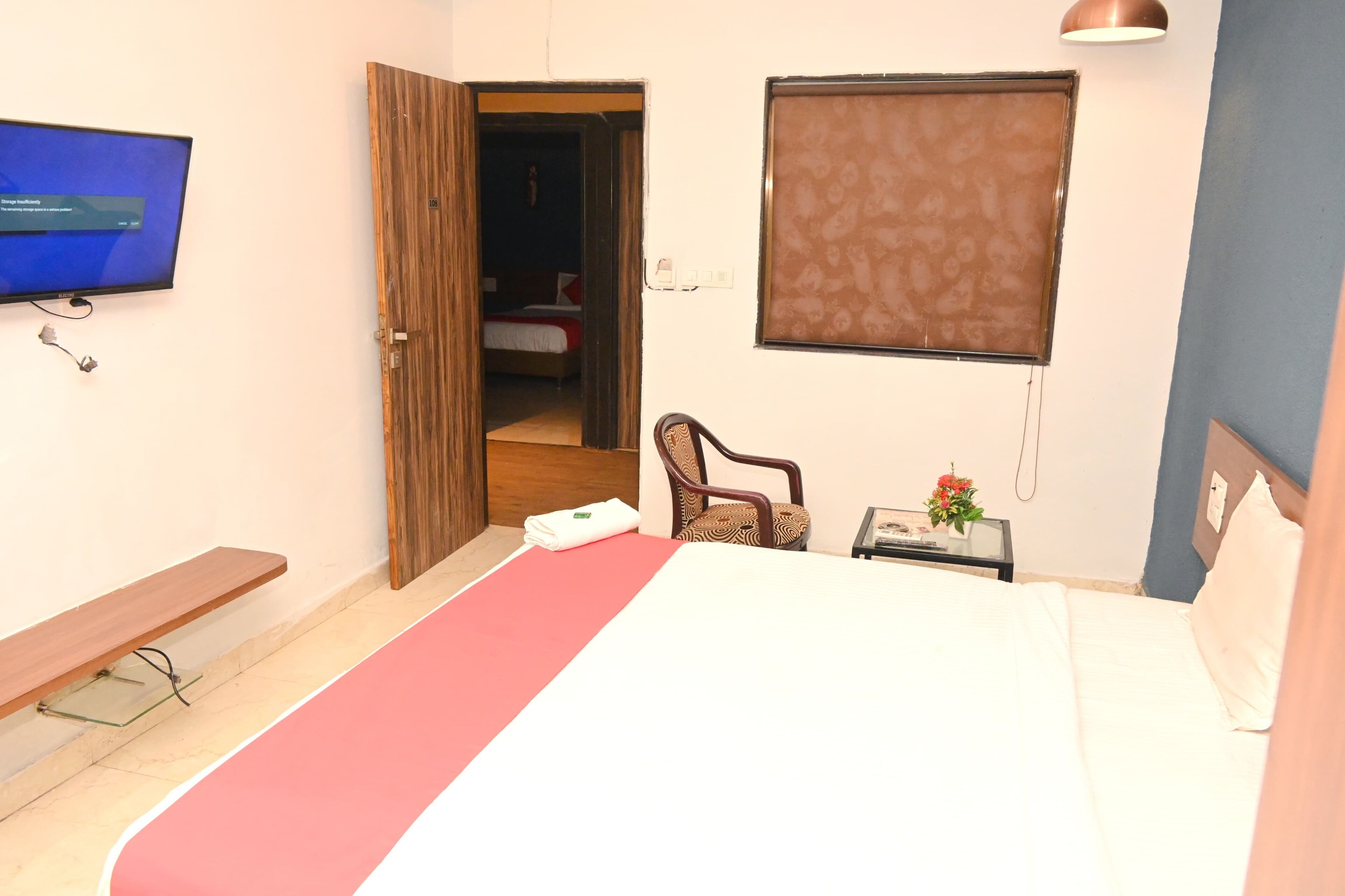 A hotel room with a bed, a chair, a small table with a flower, a TV, and a window with a blind.