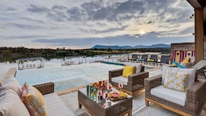 A poolside area with wicker furniture, a glass table with drinks, and a view of a lake and mountains. - Sunset Oaks Estate I: Waterfront, Infinity Pool, Hot Tub & Game Room! (Kingsland)
