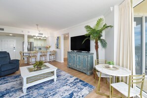 A modern living room with a white coffee table, blue patterned rug, a dining area with a round table, and a kitchen in the background.