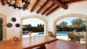 A wooden dining table with a bowl of oranges and two wine glasses, set against a backdrop of a swimming pool and outdoor seating area. - LES PALMERES (LLEIDA). Quite and peaceful (Pollensa)