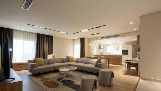 A modern living room with a sectional sofa, a coffee table, and ottomans. There is a dining area with a table and chairs in the background. - Kennah Milena | Akoya Residence, New Cairo (New Cairo)