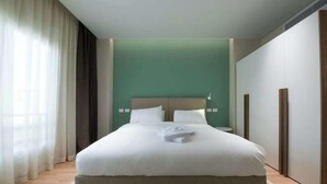A neatly made bed with white linens and a folded towel, a bedside table with a lamp, and a green accent wall. - Kennah Milena | Akoya Residence, New Cairo (New Cairo)