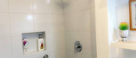 Une douche équipée d’un pommeau de douche à main, d’une barre de douche à main, et d’une étagère sur laquelle sont posés deux flacons de shampoing Pantene et Olay.