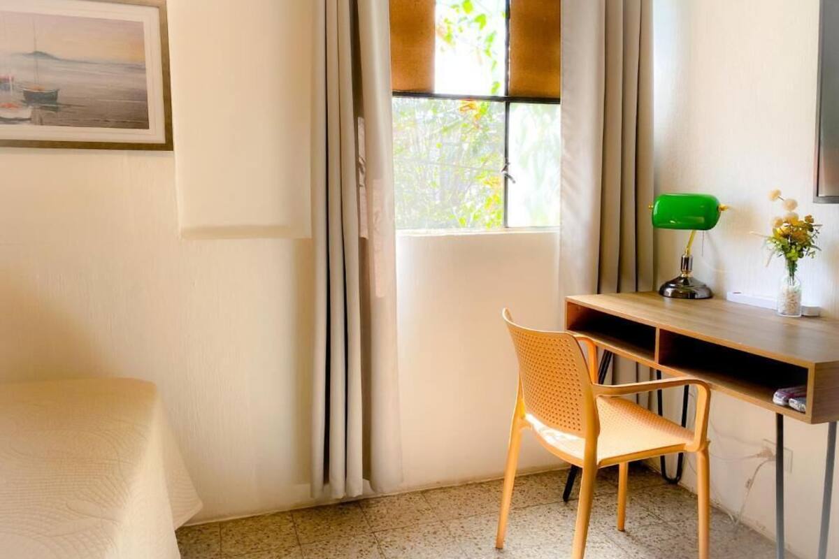 Een kamer met een bed, een bureau met stoel, een raam met gordijnen en een lijst met een afbeelding aan de muur.