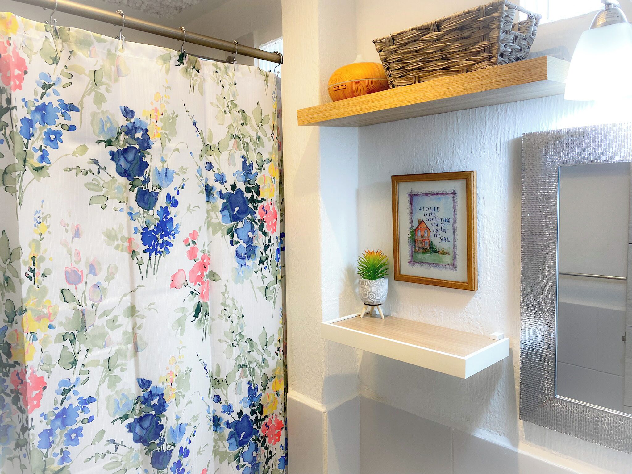 A bathroom with a floral shower curtain, a small potted plant, a framed quote on the wall, and a shelf with a basket and a lamp.