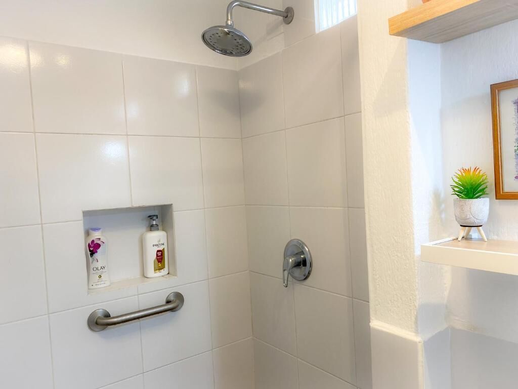 A shower area with white tiles, a handheld showerhead, a grab bar, and a shelf with toiletries.