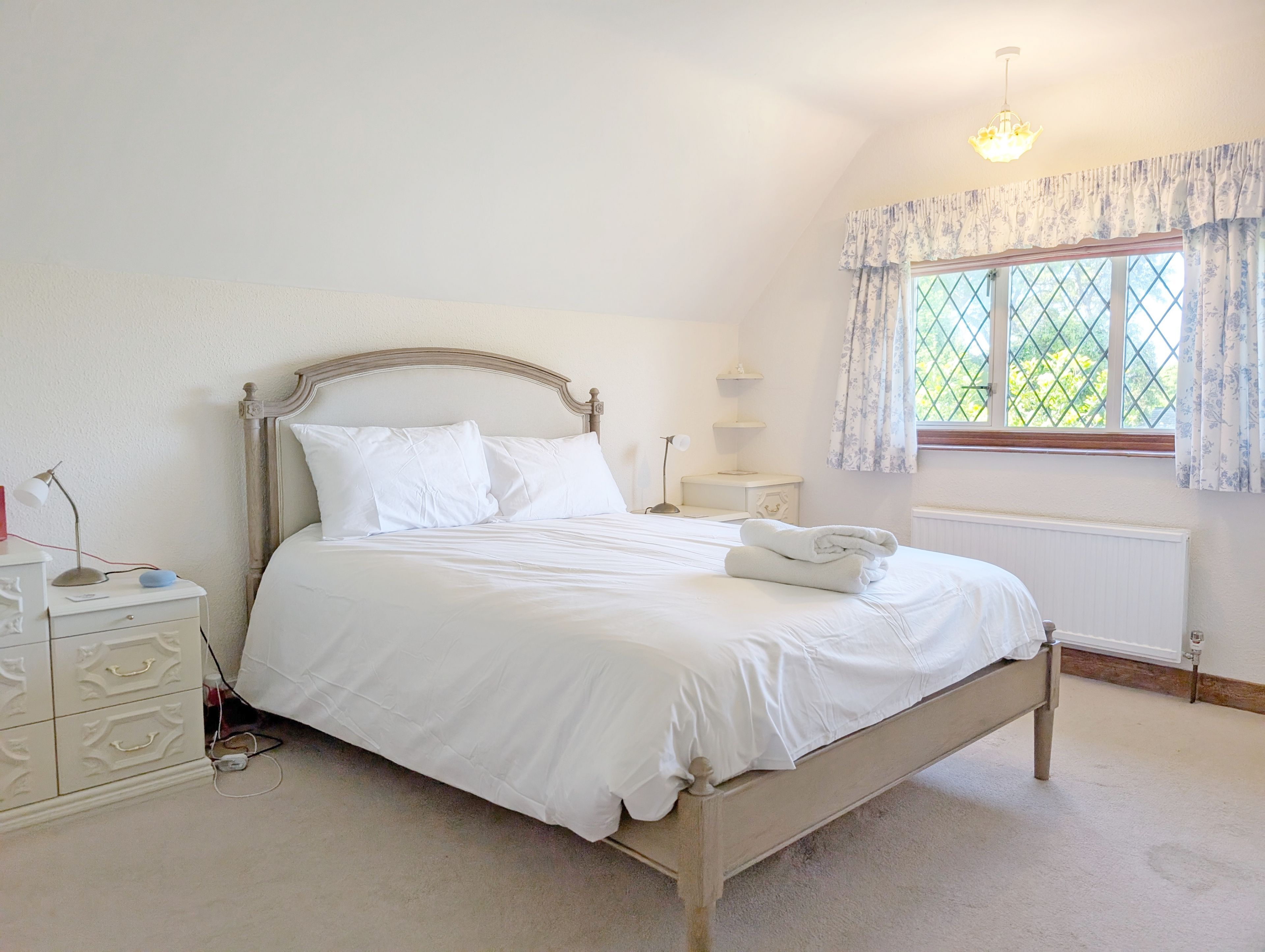 A neatly made bed in a room with a window, a bedside table, and a lamp.