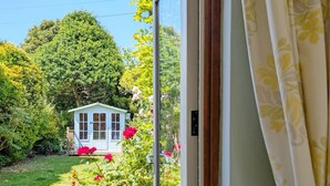 Une vue à travers la fenêtre d’une petite maison bleu clair, avec une porte blanche, entourée d’une végétation luxuriante et de fleurs rouges.