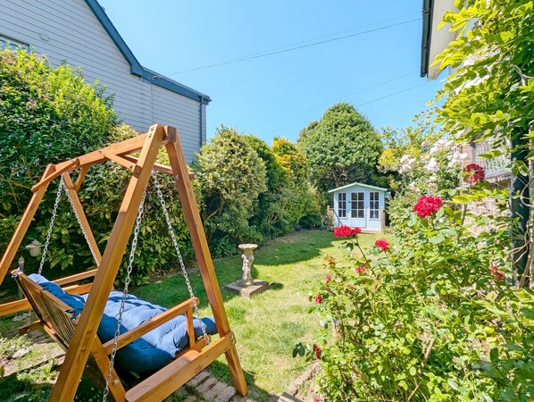Een houten schommel in een achtertuin met een kleine schuur, weelderige begroeiing en een tuin met rode bloemen.