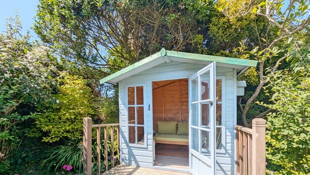 Une petite cabane en bois dotée d’un porche et d’un banc, entourée d’une végétation luxuriante.