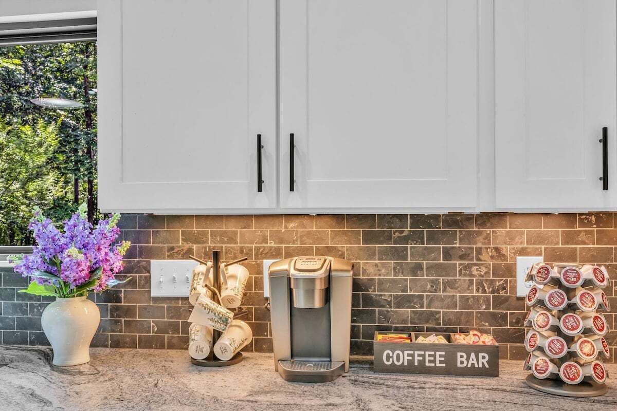 Une cuisine avec des meubles blancs, une machine à café, un panneau indiquant le coin café, et une pile de tasses à café.