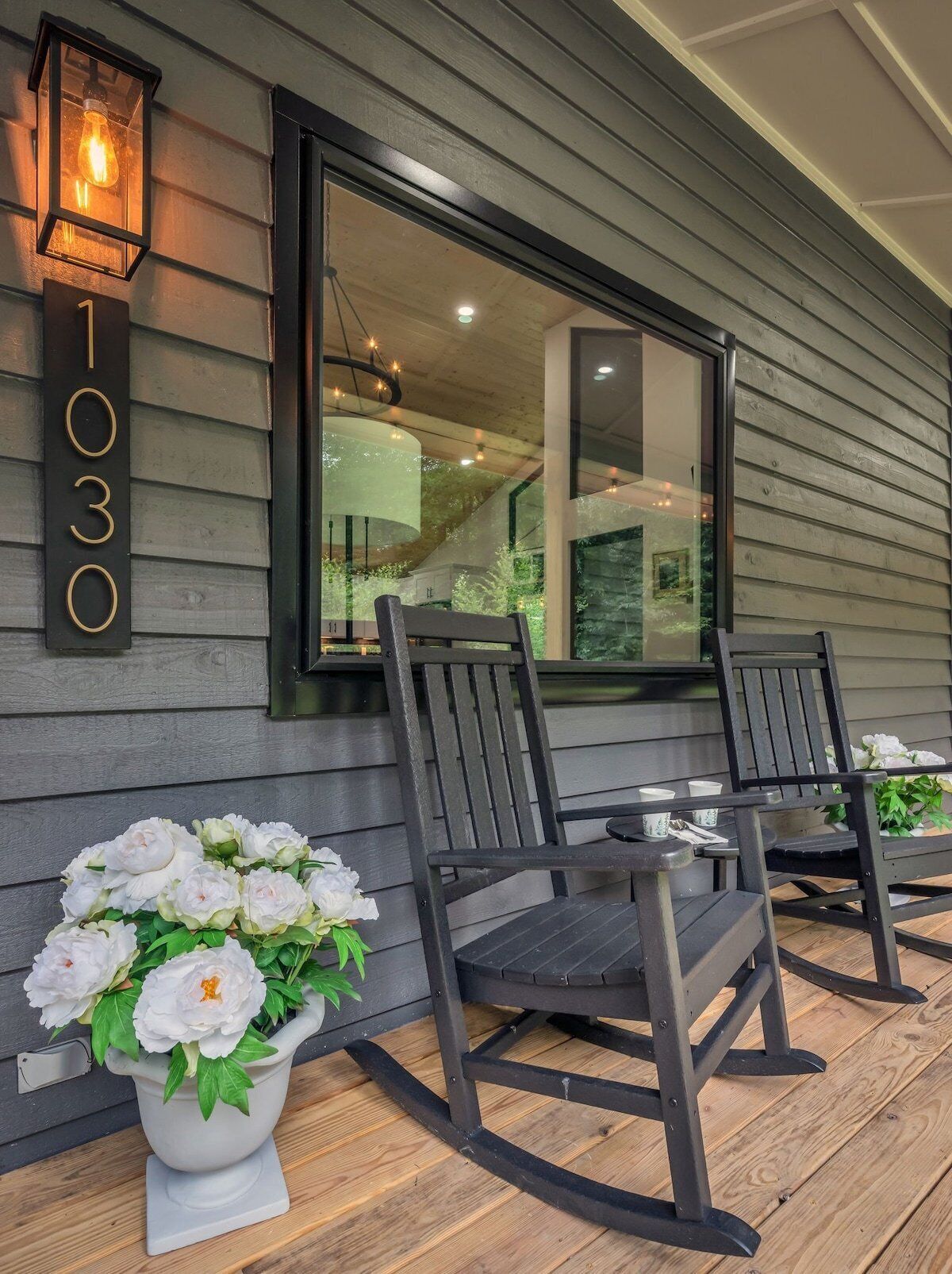 A porch with two rocking chairs, a table, and a potted plant with white flowers. A large window reflects the interior of the house.
