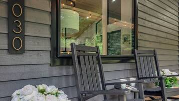 A porch with two rocking chairs, a table, and a potted plant with white flowers. A large window reflects the interior of the house.