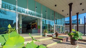 Entrada de hotel con amplios ventanales de vidrio, un patio con baldosas y plantas en macetas.
