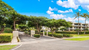 A gated entrance to a residential area with lush greenery, a clear blue sky, and a multi-story building with balconies. - Big Island Vista Waikoloa B203 2 Bedroom Condo (Waikoloa)