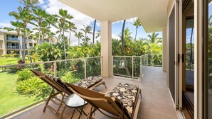 A balcony with two lounge chairs and a table, overlooking a lush garden and palm trees. - Big Island Vista Waikoloa B203 2 Bedroom Condo (Waikoloa)