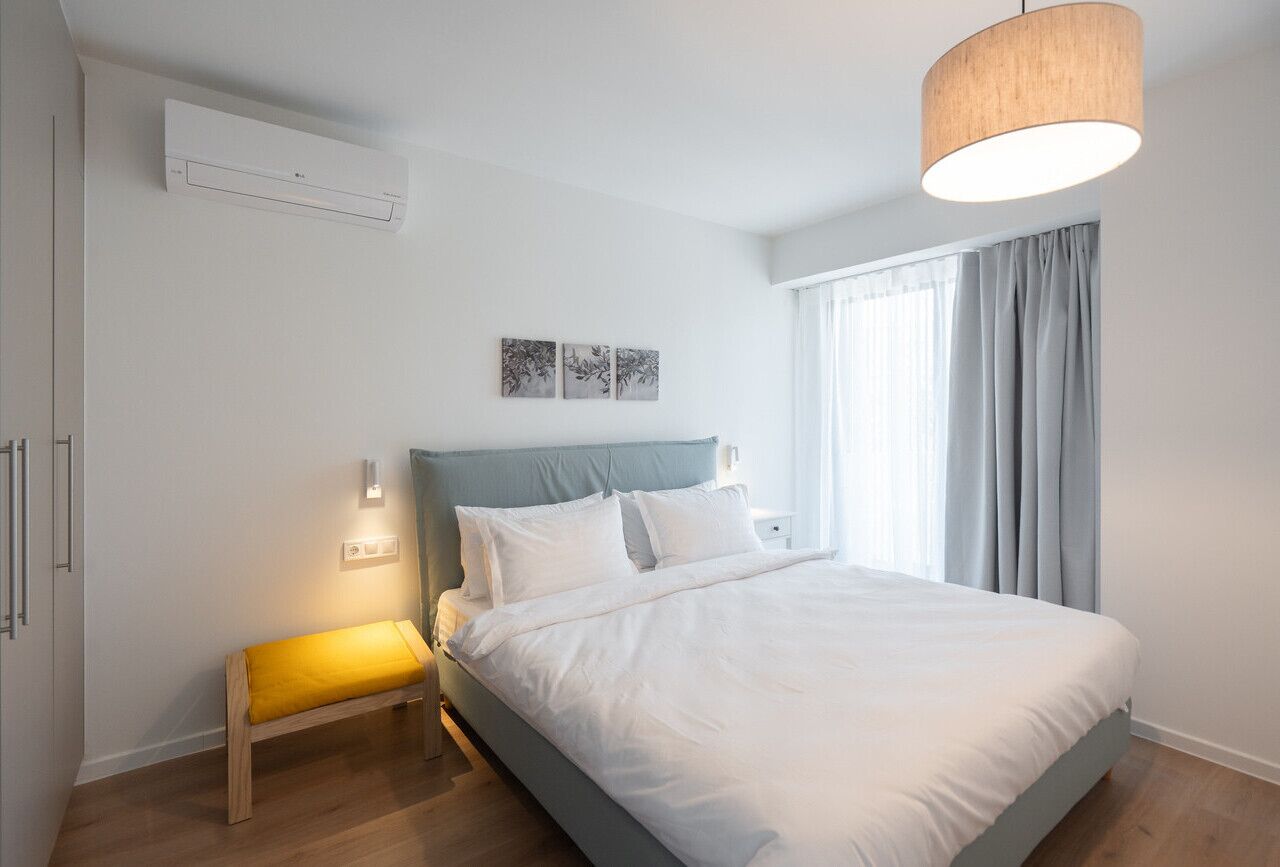 Quarto com cama, mesa de cabeceira, banco, ar condicionado fixado na parede e janela com cortinas.
