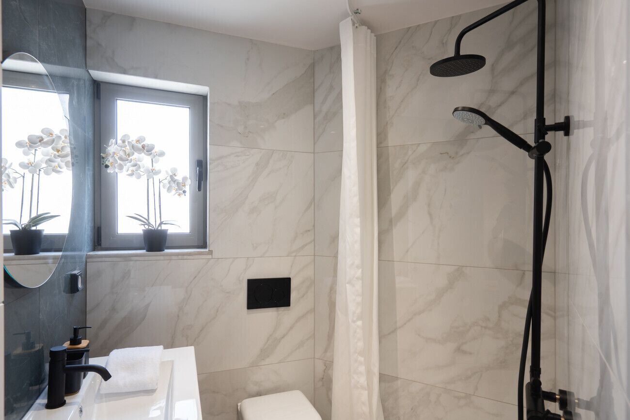 A modern bathroom with a white marble shower, a large mirror, and a window with potted plants.