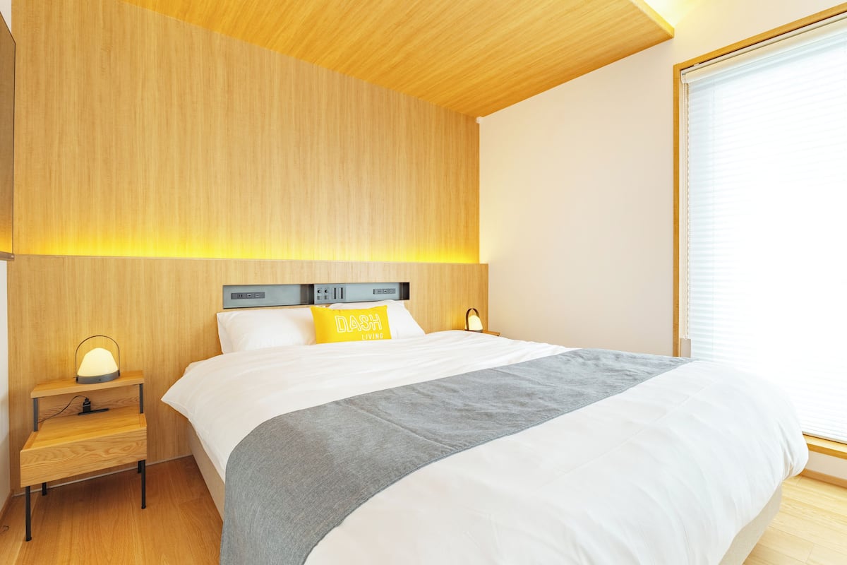 A modern hotel room with a wooden headboard, a bedside table with a lamp, a bed with white bedding and a gray throw, and a window with blinds.
