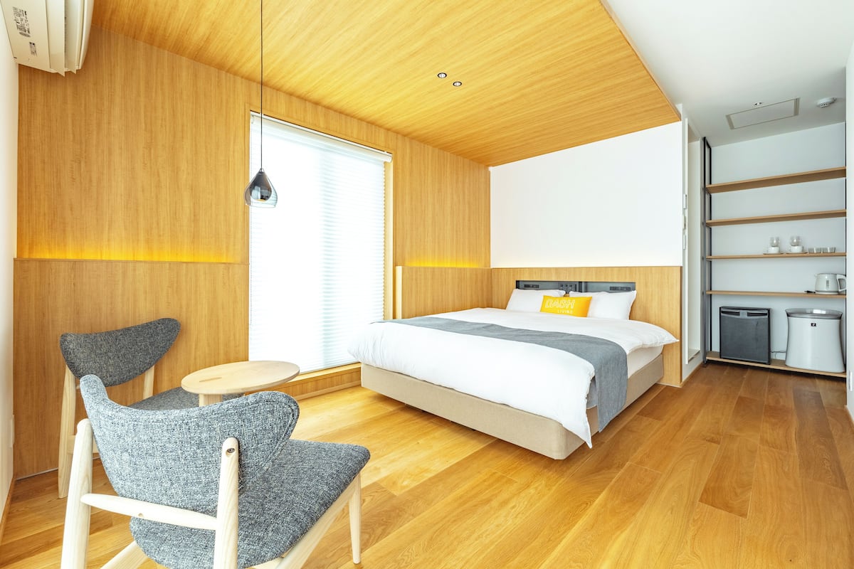 A modern hotel room with a wooden ceiling, a bed with a grey and white duvet, a grey upholstered chair, a small round table, and a built-in wardrobe with shelves.