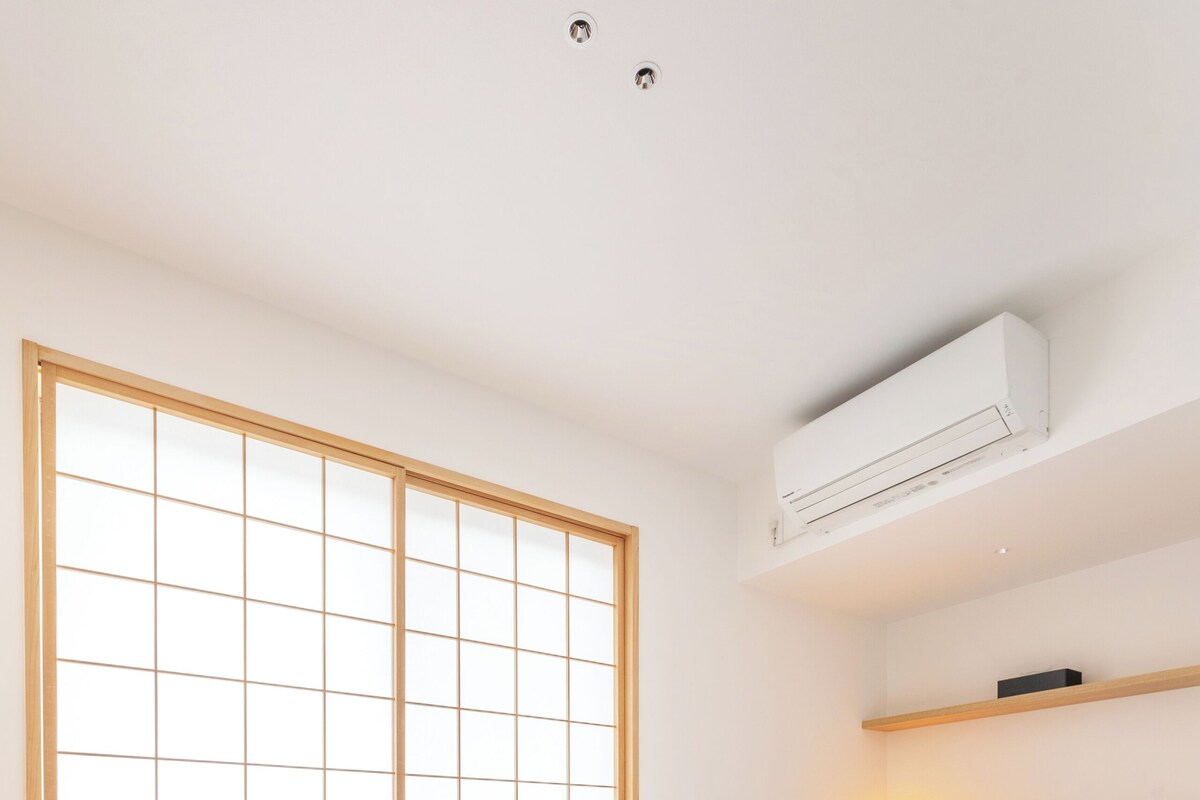 A bedroom with a bed, a desk, a chair, a window with a grid pattern, and an air conditioning unit.