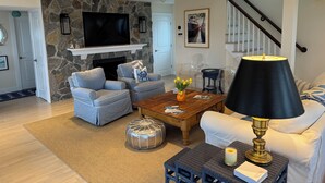 A living room with a stone wall, a fireplace, a flat-screen TV, grey sofas, a coffee table, and a staircase. - 4BR Oceanview Academic Rental in Stonington CT 09/01 thru 05/31 (Stonington)
