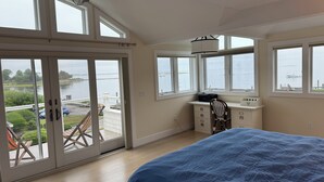 A bedroom with a large bed, a desk with a chair, and a view of the water through large windows. - 4BR Oceanview Academic Rental in Stonington CT 09/01 thru 05/31 (Stonington)