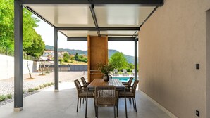 Un área de comedor al aire libre techada con vista a la piscina, que incluye una mesa y sillas, y vistas del paisaje circundante.