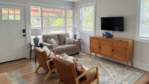Ein Wohnzimmer mit einer Couch, zwei Sesseln, einem Holzschrank, einem Fernseher und großen Fenstern mit Jalousien.
