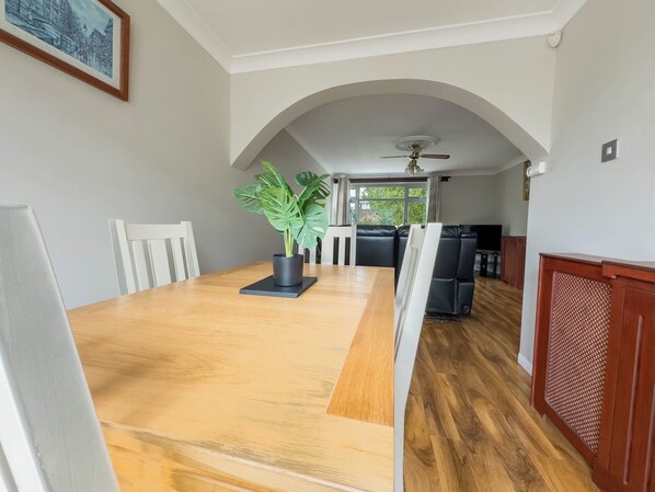 A dining area with a wooden table, white chairs, a potted plant, and a view of the living room through an arched doorway.