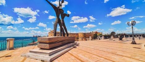 A plaza with a prominent sculpture, a clear blue sky, and a coastal backdrop.
