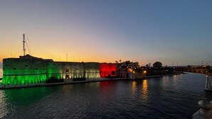 Blick auf das Wasser mit einer historischen Festung, deren Bereiche rot und grün beleuchtet sind, einem ruhigen Fluss und einer entfernteren Stadtlandschaft im Abenddämmerung.