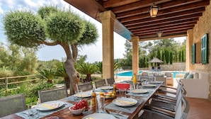 Un área de comedor al aire libre con una piscina al fondo, rodeada de follaje y muros de piedra.