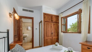 Un dormitorio con cama, armario de madera, ventana con cortinas, mesita de noche y baño visible a través de una puerta abierta.