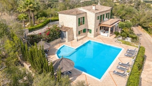 Una gran piscina rectangular con solárium de piedra, rodeada por una casa de piedra con contraventanas verdes, techo de paja y vistas al campo.