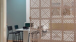 Een kamer met een wit bureau, twee zwarte stoelen, een computermonitor en een decoratieve scheidingswand met een geometrisch patroon.