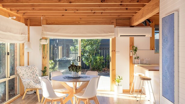 A modern kitchen with wooden beams, a dining area with white chairs, and a large window overlooking a garden.