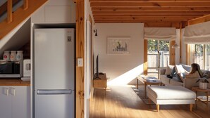 A modern kitchen with a refrigerator, microwave, and cabinets. There is a dining area with a table and chairs, and a living room with a sofa and a plant.