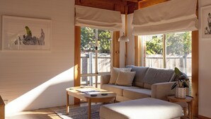 A cozy living room with a sofa, ottoman, and coffee table, large windows with blinds, and a framed artwork on the wall.