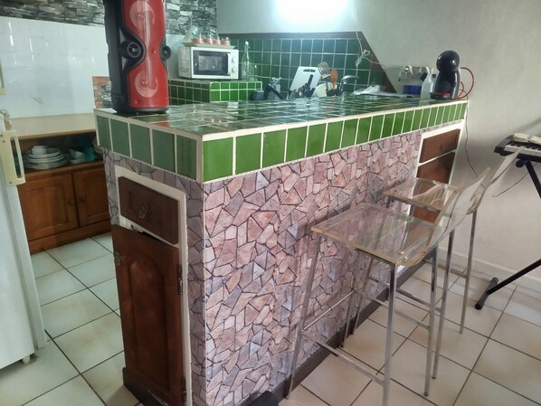 Uma cozinha com painel de azulejos verde e rosa, micro-ondas, eletrodoméstico vermelho e um espaço de bar com uma cadeira.