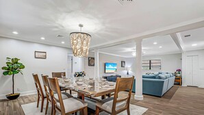 Un espace repas moderne avec une table en bois, des chaises et un lustre. On aperçoit un canapé et un téléviseur à l’arrière-plan.