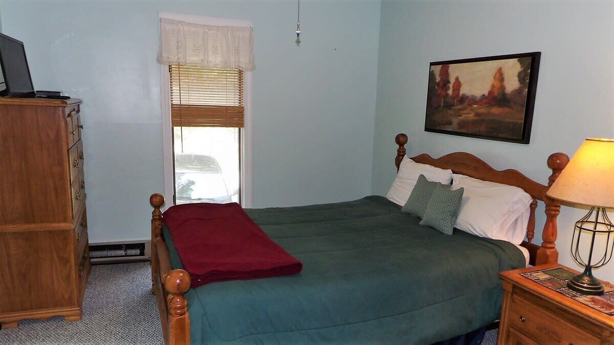 A bedroom with a bed, a wooden nightstand with a lamp, a dresser, a window with blinds, and a painting on the wall.