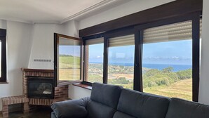 A living room with a fireplace, large windows, and a view of the sea. - La Atalaya del Cabo - 4800 (Arnuero)