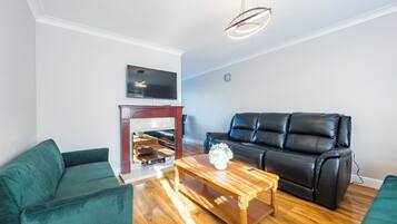 A modern living room with a black leather sofa, a green armchair, and a wooden coffee table. A flat-screen TV is mounted on the wall above a fireplace.