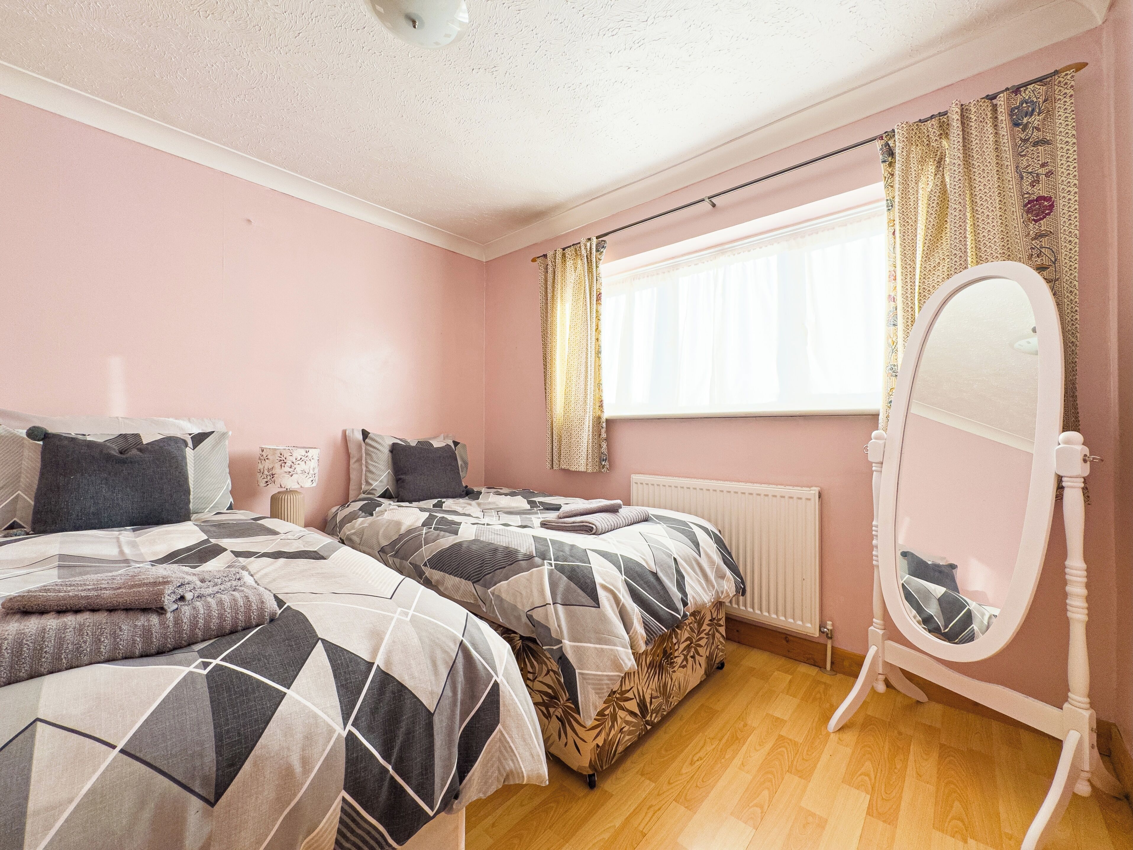 A bedroom with two beds, a radiator, a window with curtains, and a mirror.