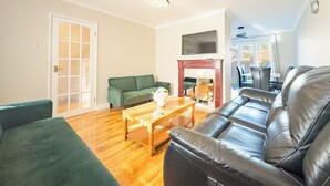 Un vaste salon comprenant un canapé vert, un divan en cuir noir et une table basse en bois. On aperçoit une cheminée au fond de la pièce, et une porte donne accès à une autre pièce.