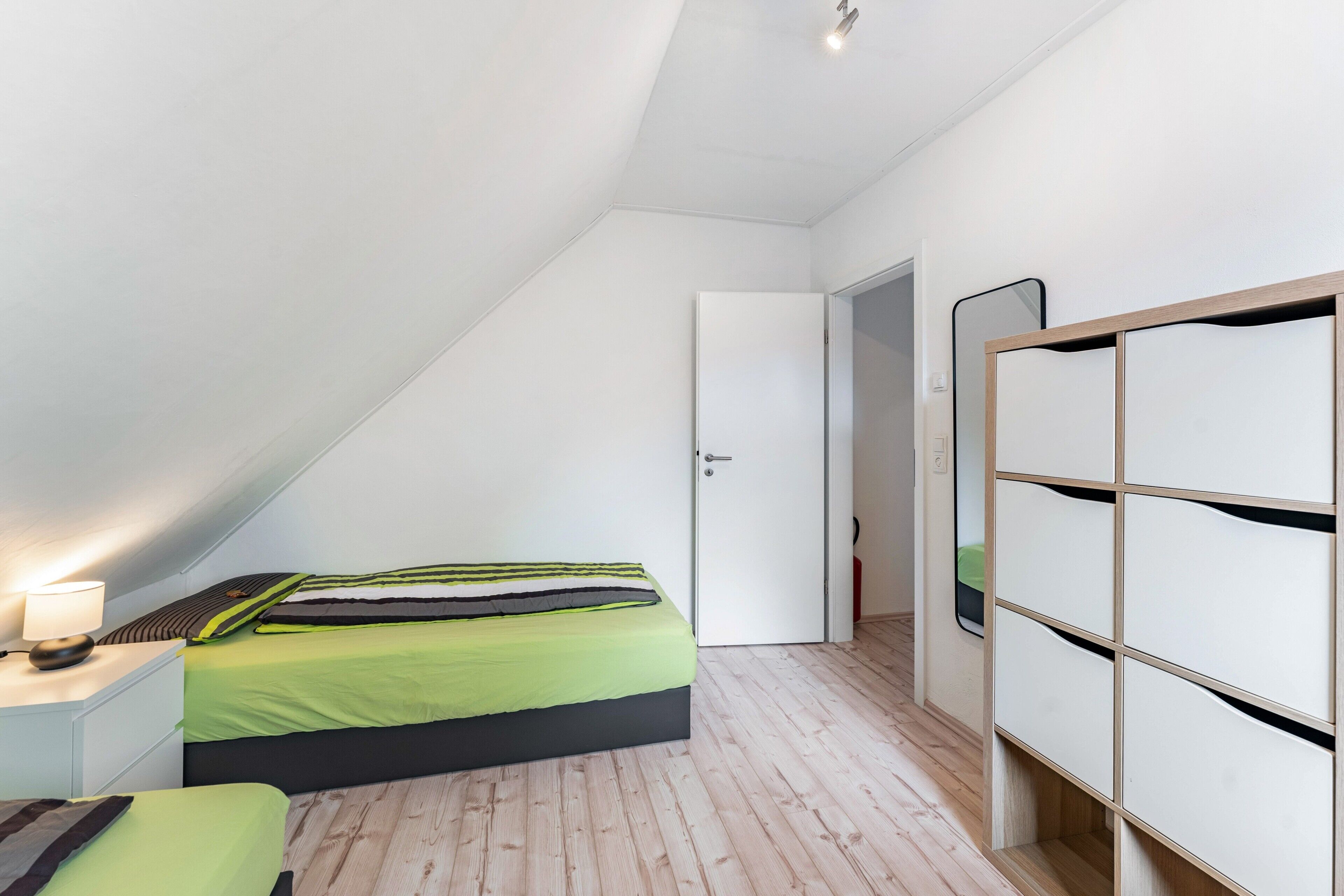Quarto com cama, mesa de cabeceira com luminária, armário e porta para outro cômodo.